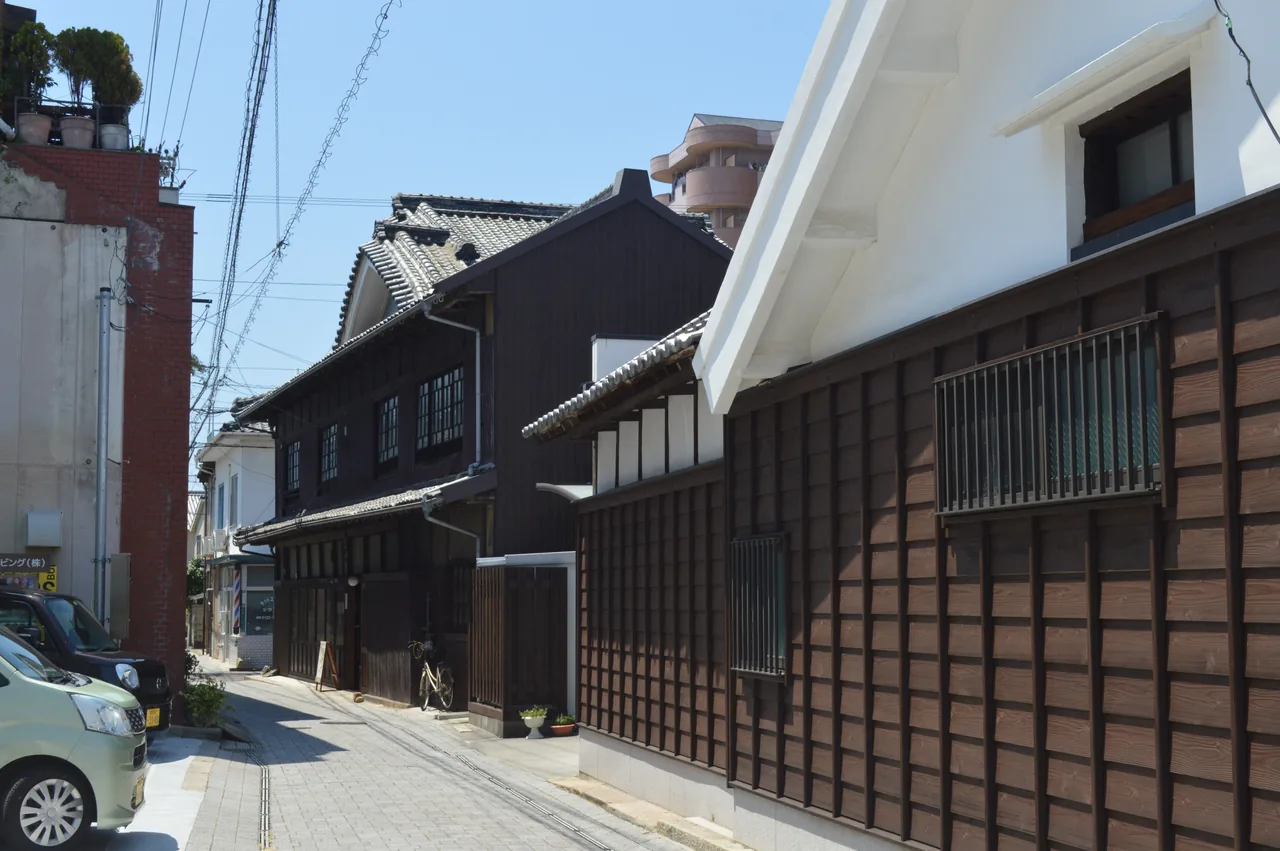 A narrow sunny lane in Mitsuhama lined with dark wooden two-storey merchant houses and a white plaster storehouse, one bicycle and no people