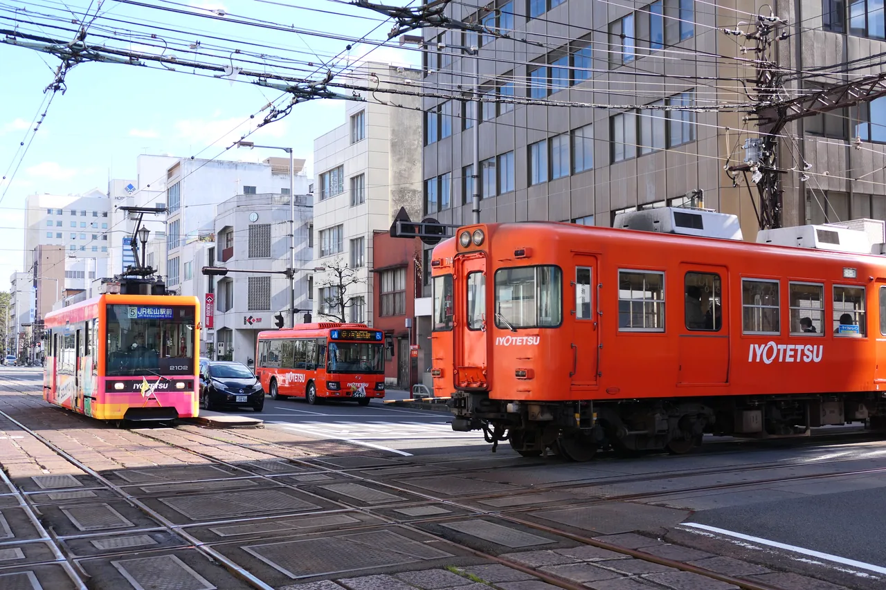 An orange suburban train crossing tram tracks at an intersection while a tram and a bus wait