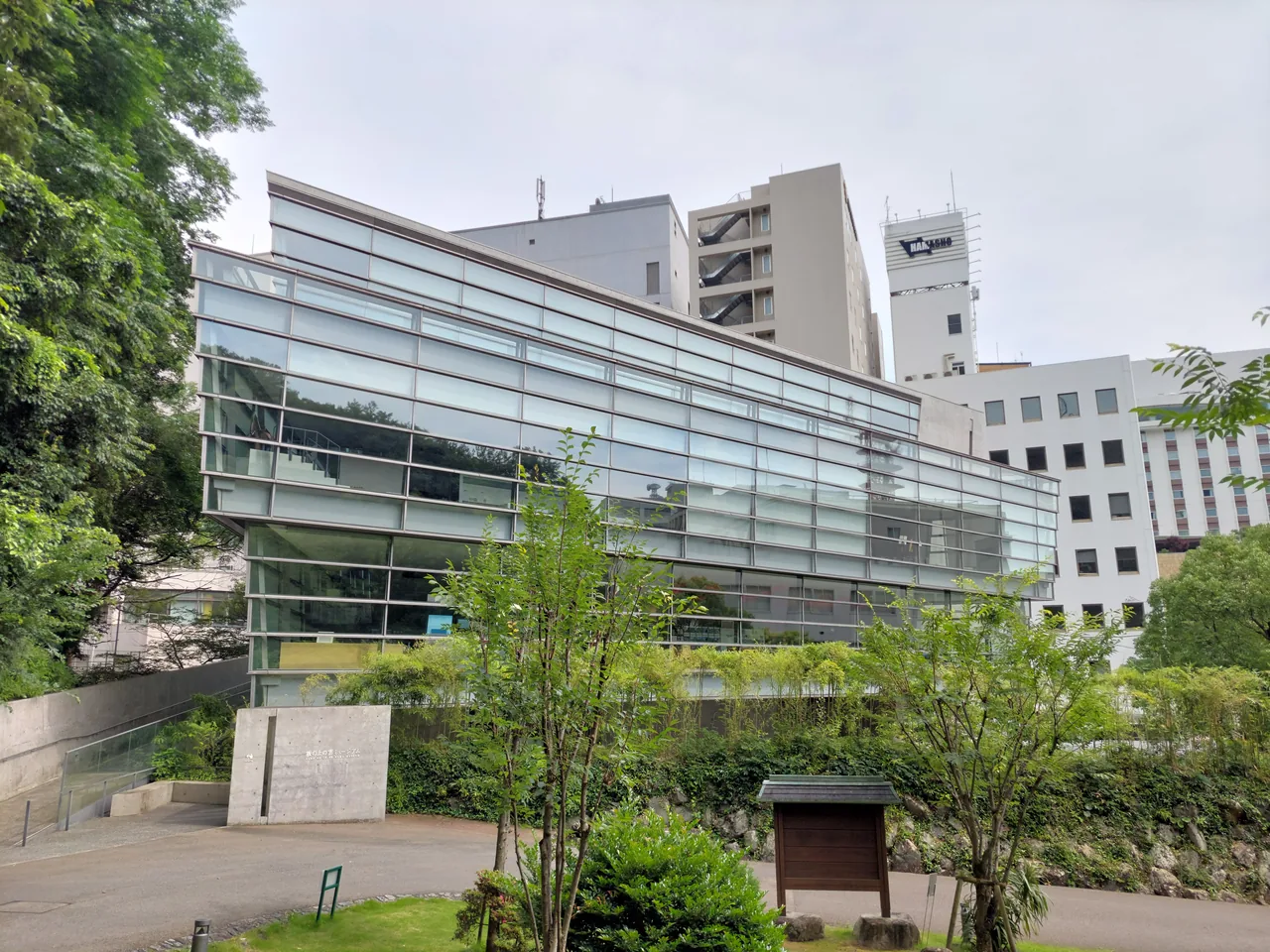 The slanted glass facade of the Saka no Ue no Kumo Museum with trees in front